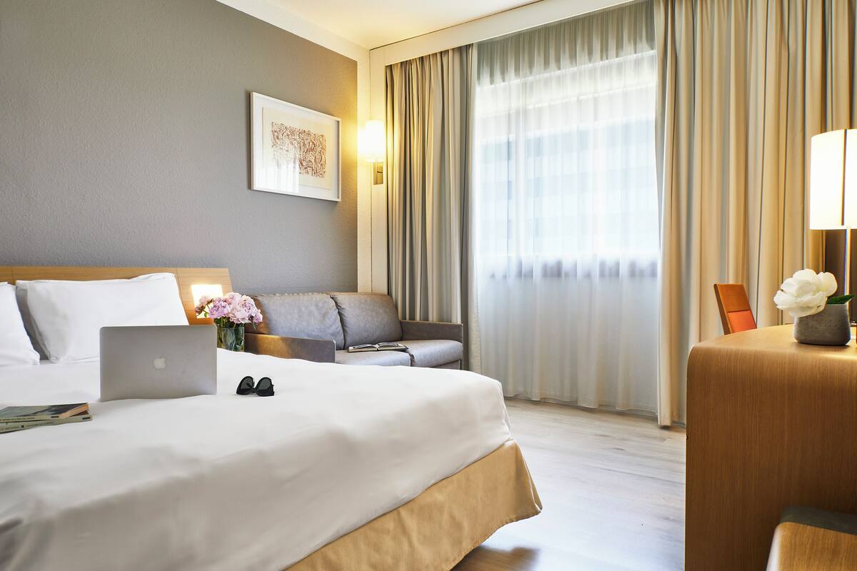 Ein Hotelzimmer mit einem Bett, einem Laptop, einer Brille, einem Sofa und einem Schreibtisch mit Lampe und Blumen.