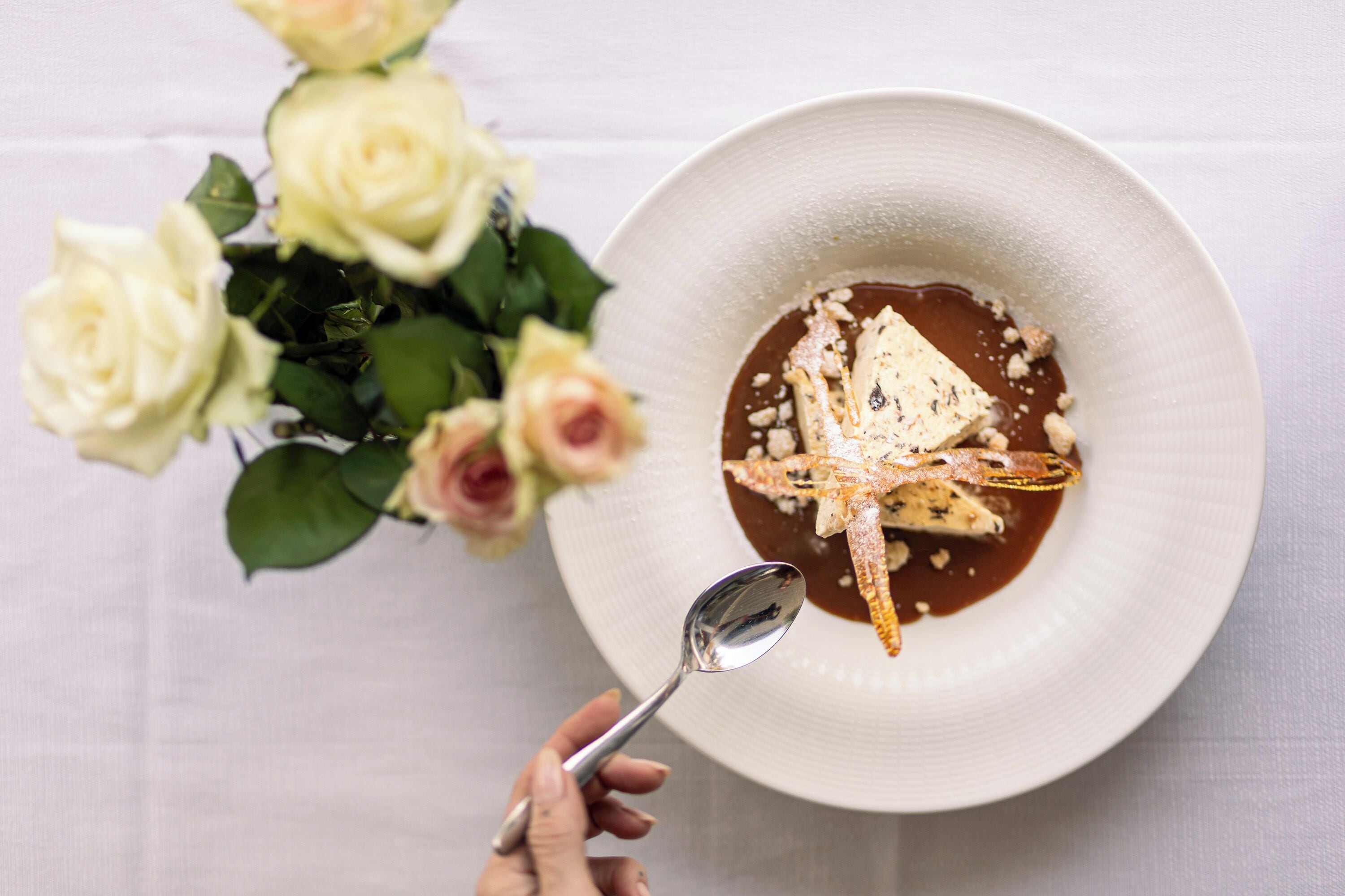Un plato blanco con un postre, una cuchara y un ramo de rosas blancas.