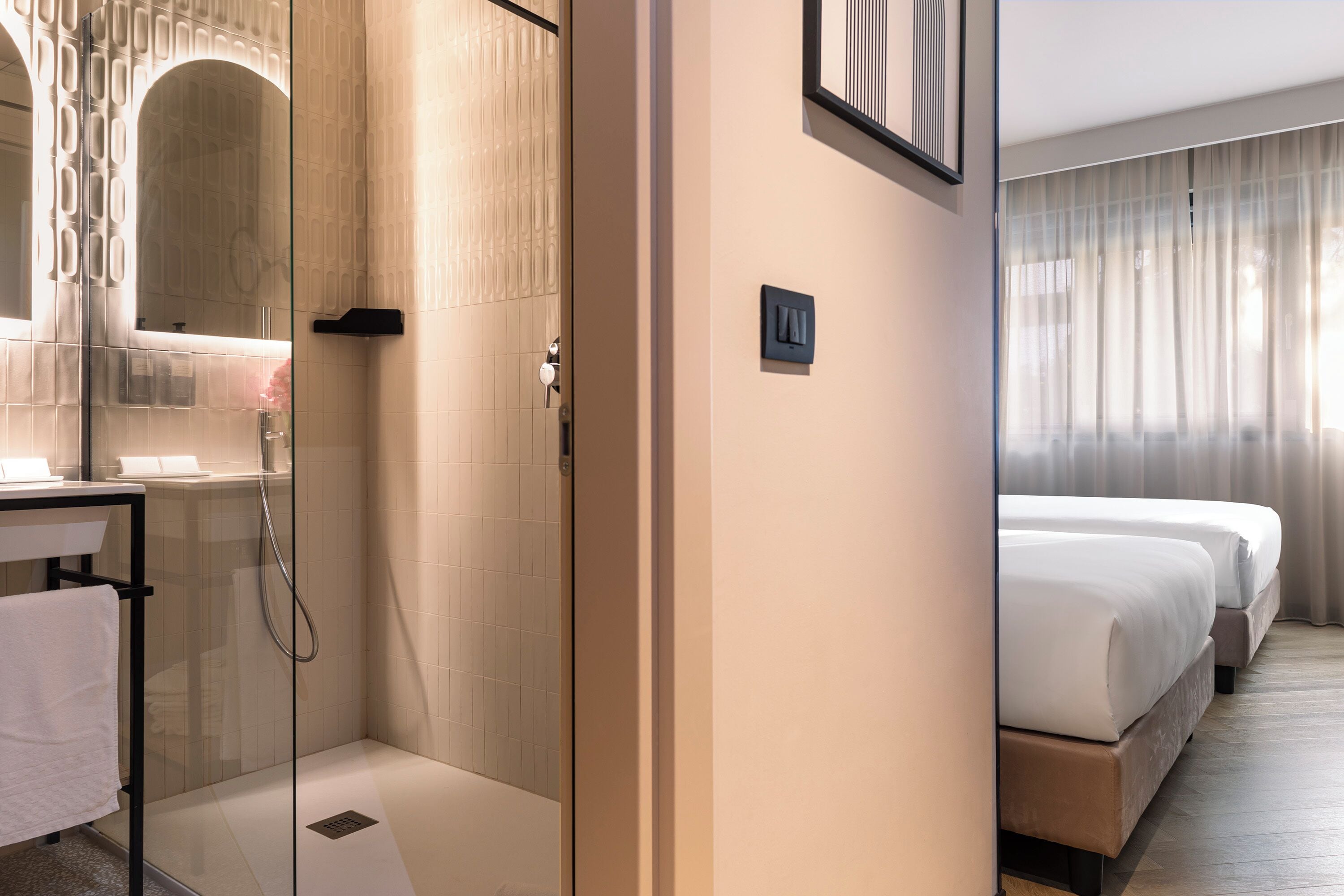 Habitación de hotel con ducha envuelta en vidrio, mesita de noche con toalla, cama con ropa blanca y ventana grande con cortinas.