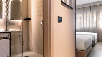 Camera d'albergo con cabina doccia in vetro, comodino con asciugamano, letto con biancheria bianca e un'ampia finestra con tende.