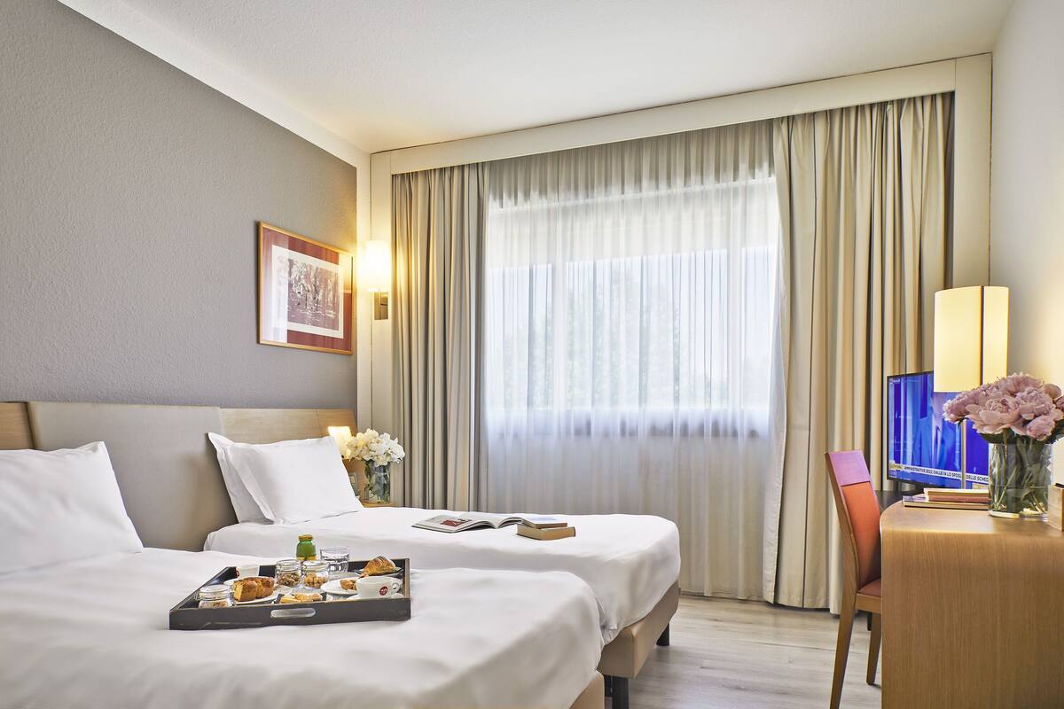 Ein Hotelzimmer mit zwei Betten, einem Schreibtisch mit Computer und einem großen Fenster mit Vorhängen.