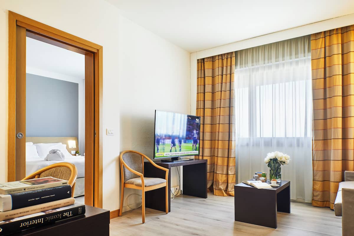 Ein Hotelzimmer mit einem Bett, einem Fernseher, einem Holzsessel, einem kleinen Tisch mit Blumenvase und einem Fenster mit Vorhängen.