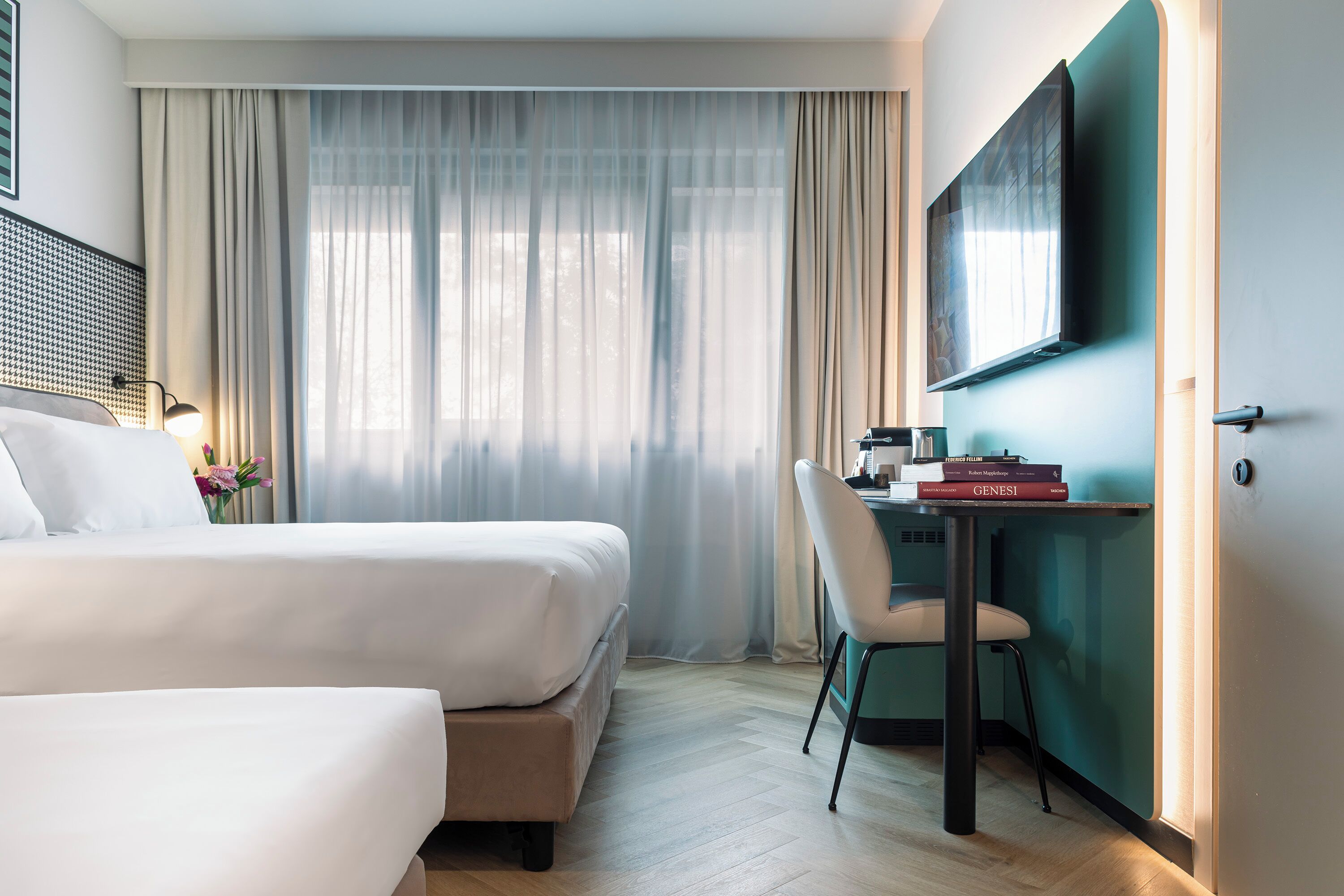 Une chambre d’hôtel avec deux lits, un bureau avec une chaise, un téléviseur fixé au mur et une grande fenêtre avec des rideaux légers.