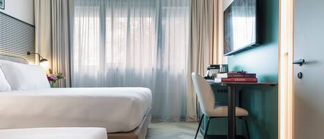 Une chambre d’hôtel avec deux lits, un bureau avec une chaise, un téléviseur fixé au mur et une grande fenêtre avec des rideaux légers.