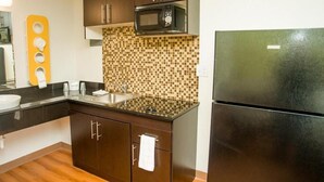 A compact kitchen with dark cabinets, a microwave, a refrigerator, a sink, and a tiled backsplash. - Motel 6 Palestine, TX (Palestine)