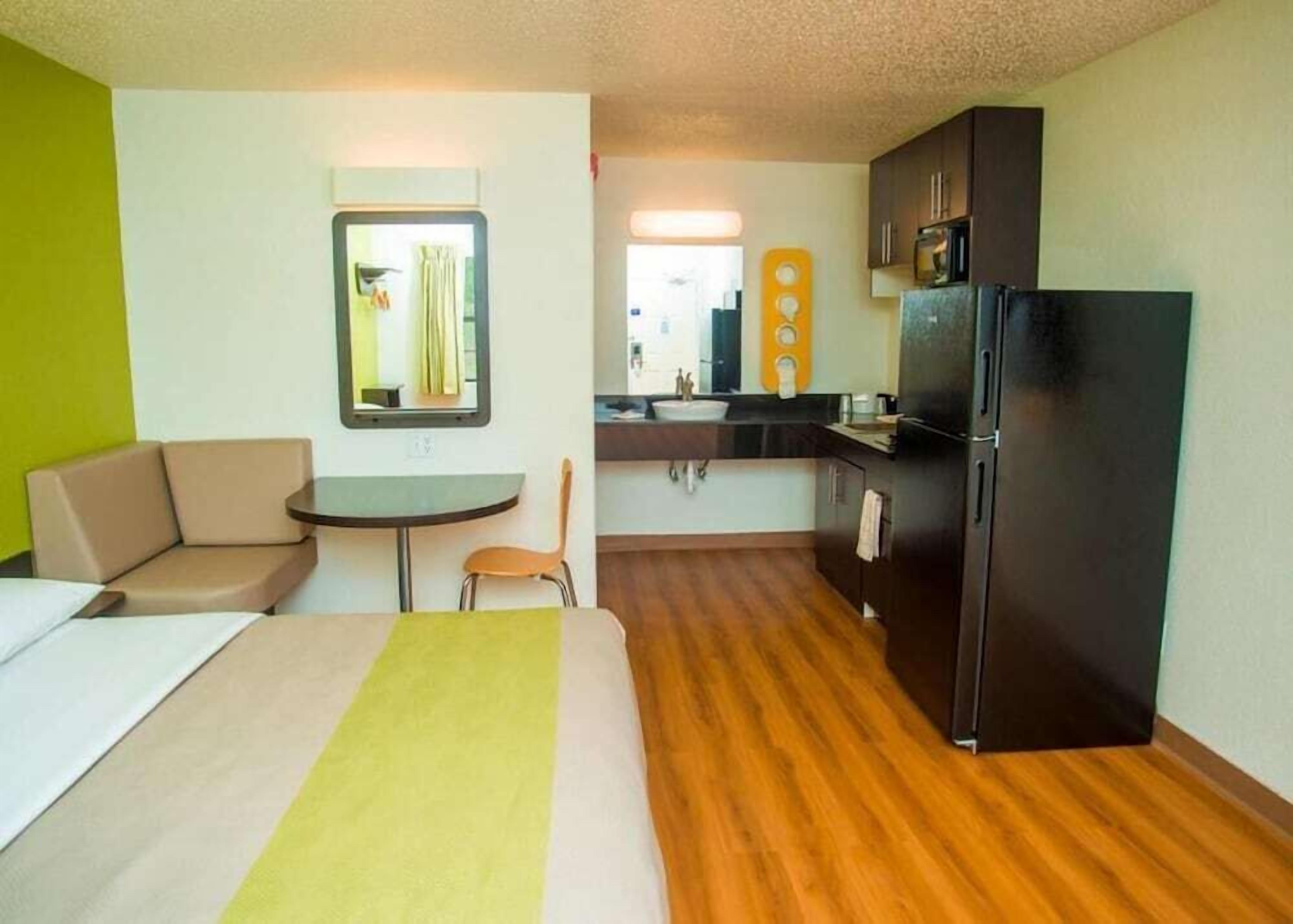 A compact hotel room with a bed, a small table, a chair, a kitchenette, and a mirror.