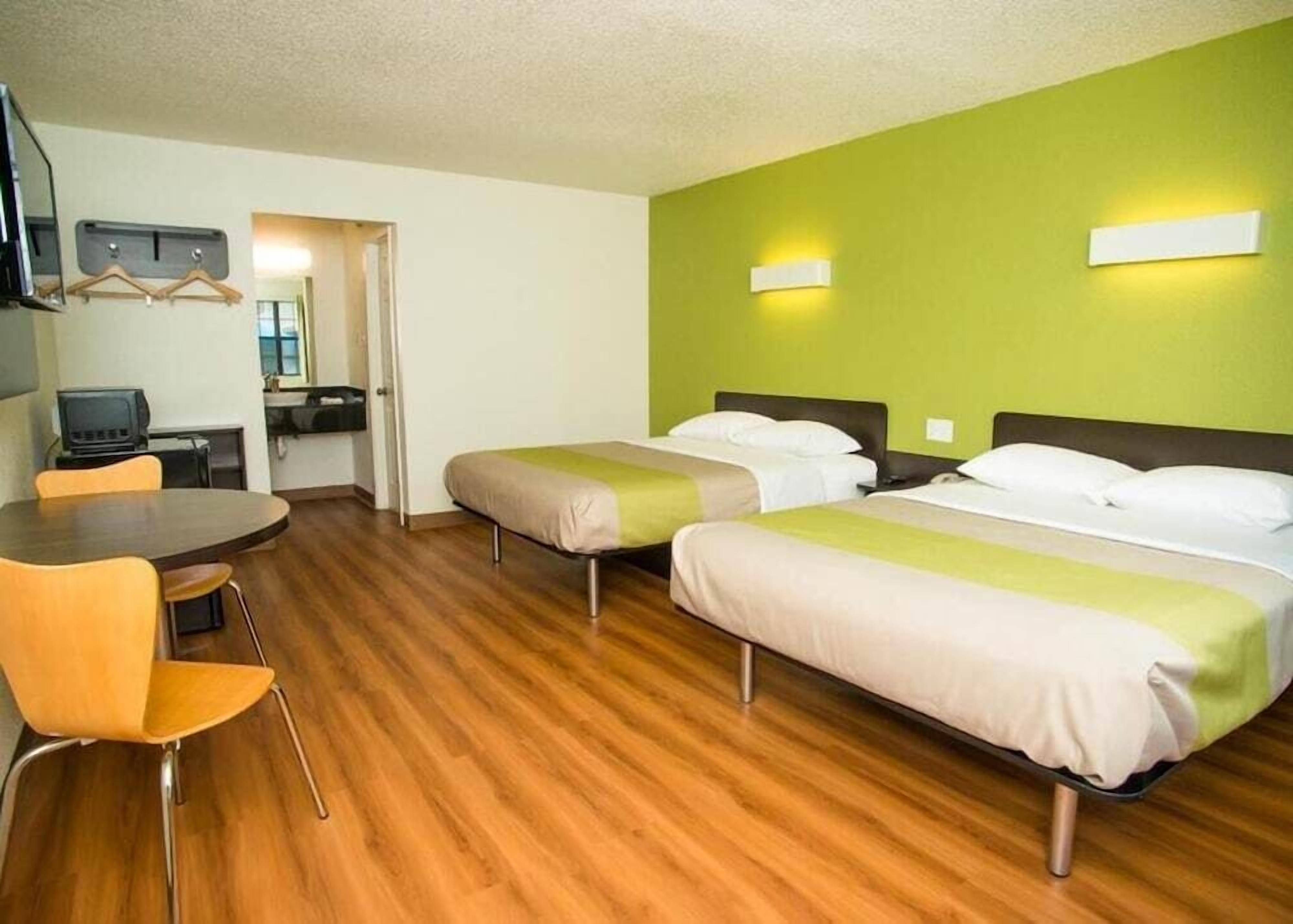 Een hotelkamer met twee eenpersoonsbedden, een houten vloer, een eettafel met stoelen, een televisie en een groene muur met twee wandlampen.