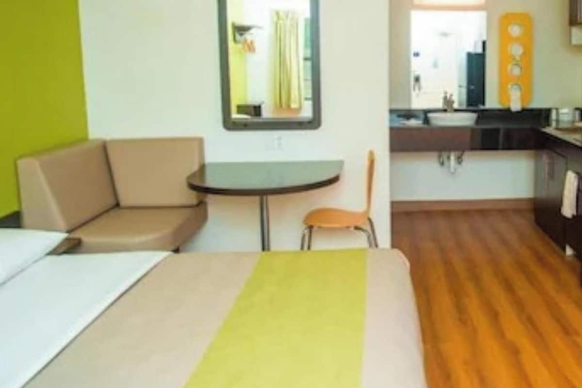 A compact hotel room with a bed, a small round table, a sofa, a chair, and a kitchenette.