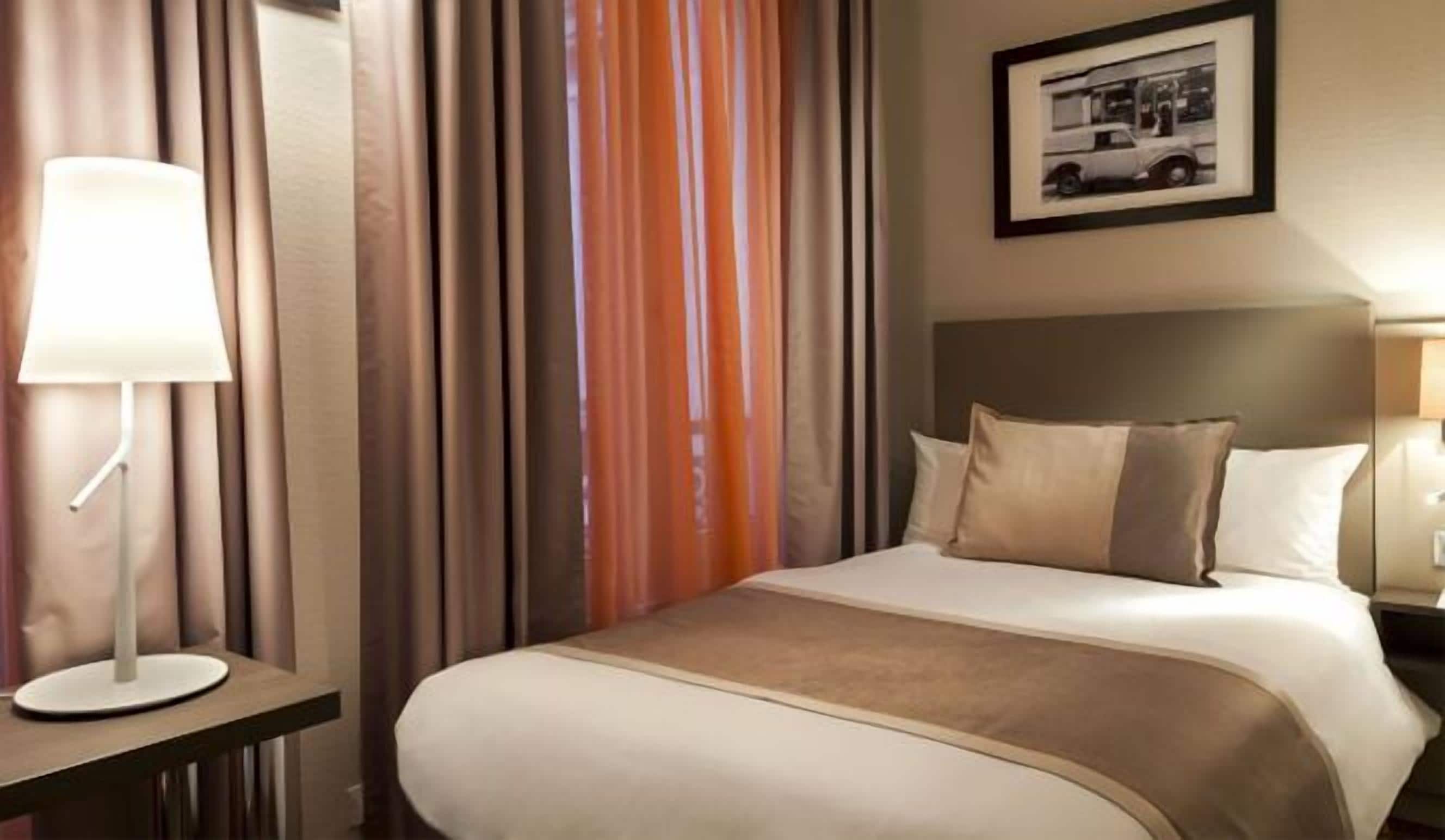 A hotel room with a bed, a nightstand, a lamp, and a framed picture on the wall.