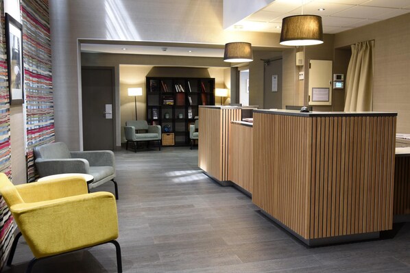 A reception area with a wooden reception desk, two chairs, a bookshelf, and a lamp.