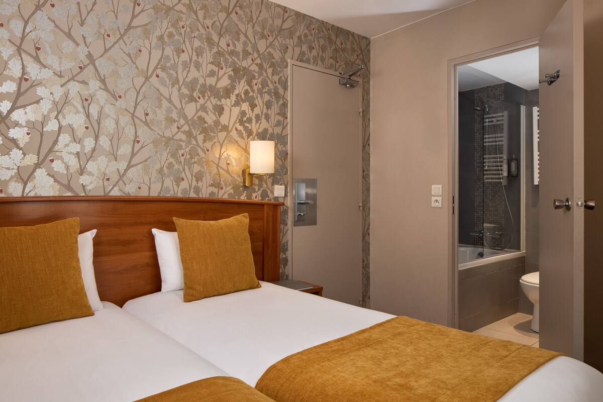 A hotel room with two beds, a wooden headboard, a wall-mounted lamp, and a bathroom visible through an open door.
