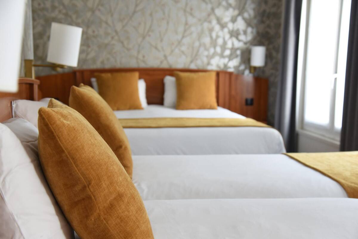 A hotel room with two beds, each with white linens and mustard-colored pillows. There is a nightstand with a lamp, and a window with curtains.