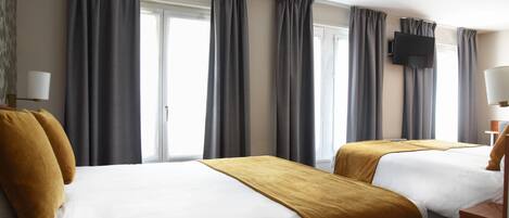 A hotel room with two beds, grey curtains, a flat-screen TV, and a lamp.