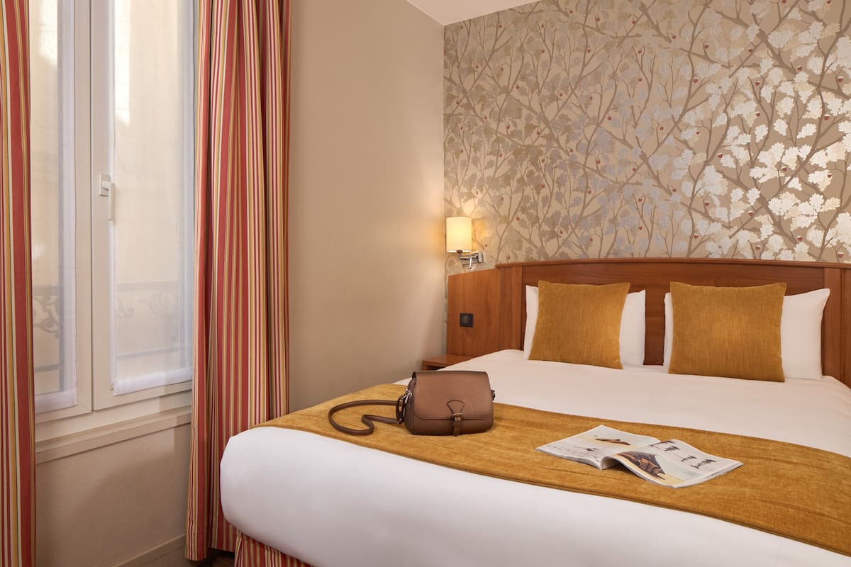A hotel room with a bed, a wooden headboard, a window with curtains, a wall with a floral pattern, and a lamp.