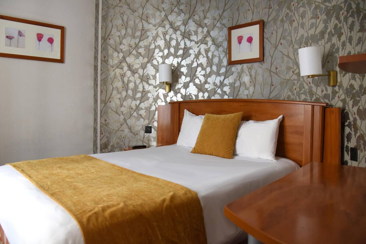 A hotel room with a bed, a wooden nightstand, a bedside table, and a wall with floral wallpaper.