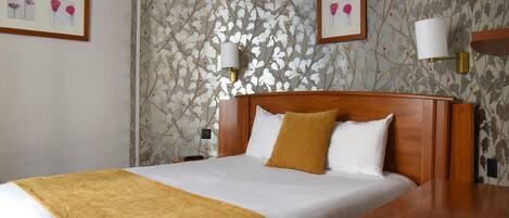 Una habitación de hotel con una cama, una mesita de noche de madera y un atril junto a la cama, y una pared con estampado floral.