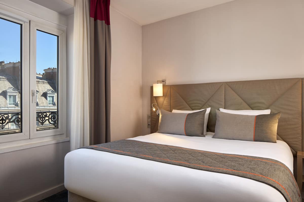 A hotel room with a large bed, a headboard with a light fixture, and a window with a view of buildings.