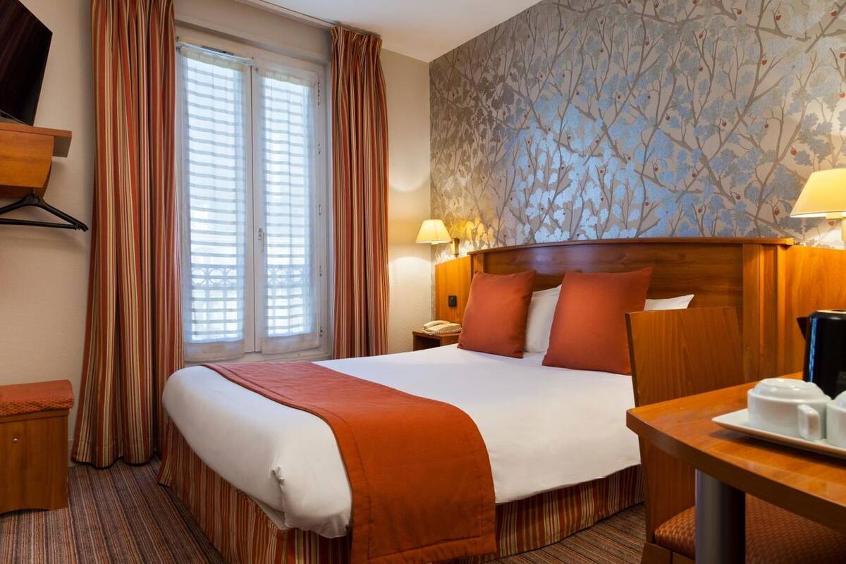 A hotel room with a bed, a desk, a chair, a nightstand, a lamp, and a window with curtains.