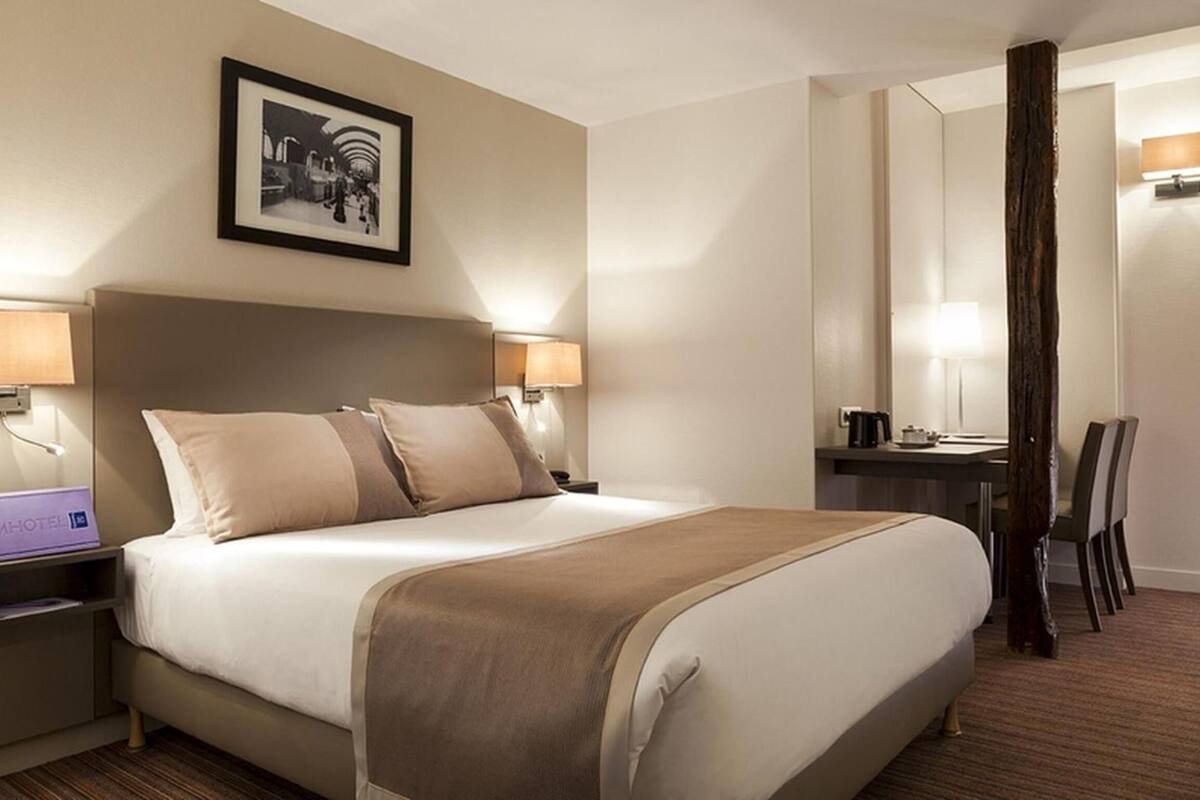 A hotel room with a bed, bedside lamps, a desk, a chair, a mirror, and a framed picture on the wall.