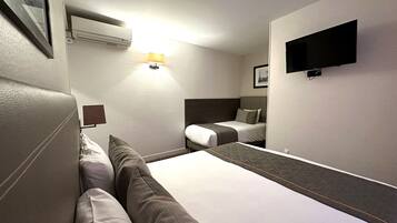 A hotel room with two beds, a wall-mounted TV, an air conditioning unit, and a bedside lamp.