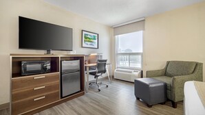 Quarto de hotel com televisão de tela plana, micro-ondas, mini-geladeira, escrivaninha com cadeira, poltrona verde, um pequeno banquinho e uma grande janela com vista.