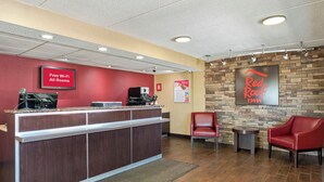 A hotel reception area with a red wall, a Red Roof Inn sign, a coffee machine, and a seating area with two red chairs. - Red Roof Inn West Monroe (West Monroe)