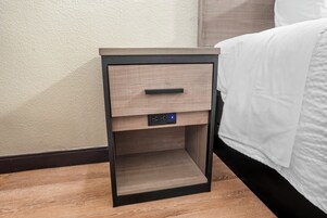 Una mesita de noche de madera con un cajón, un enchufe y un reloj digital.
