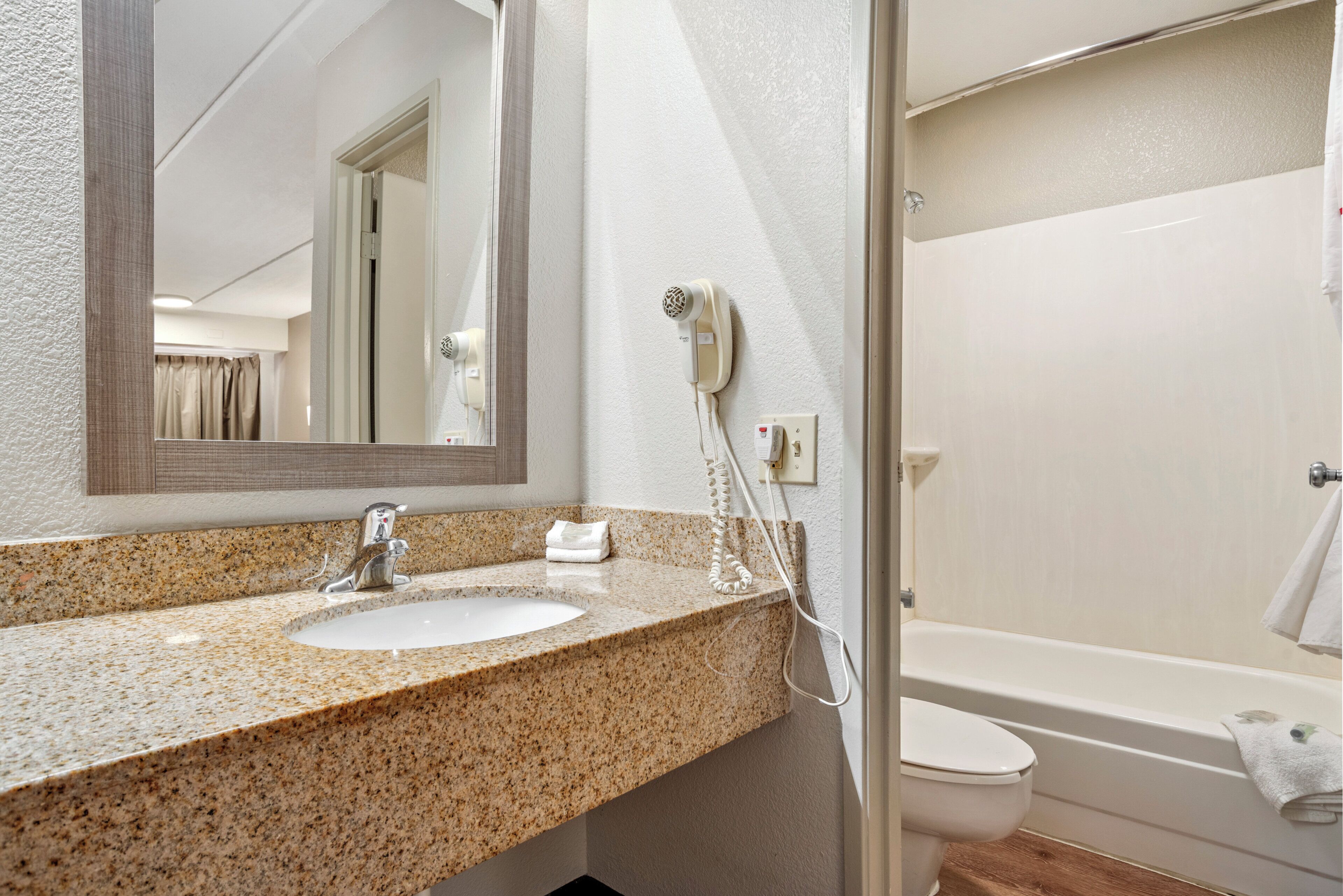 Un baño con lavamanos de encimera de granito, un espejo, un secador de pelo y una bañera.
