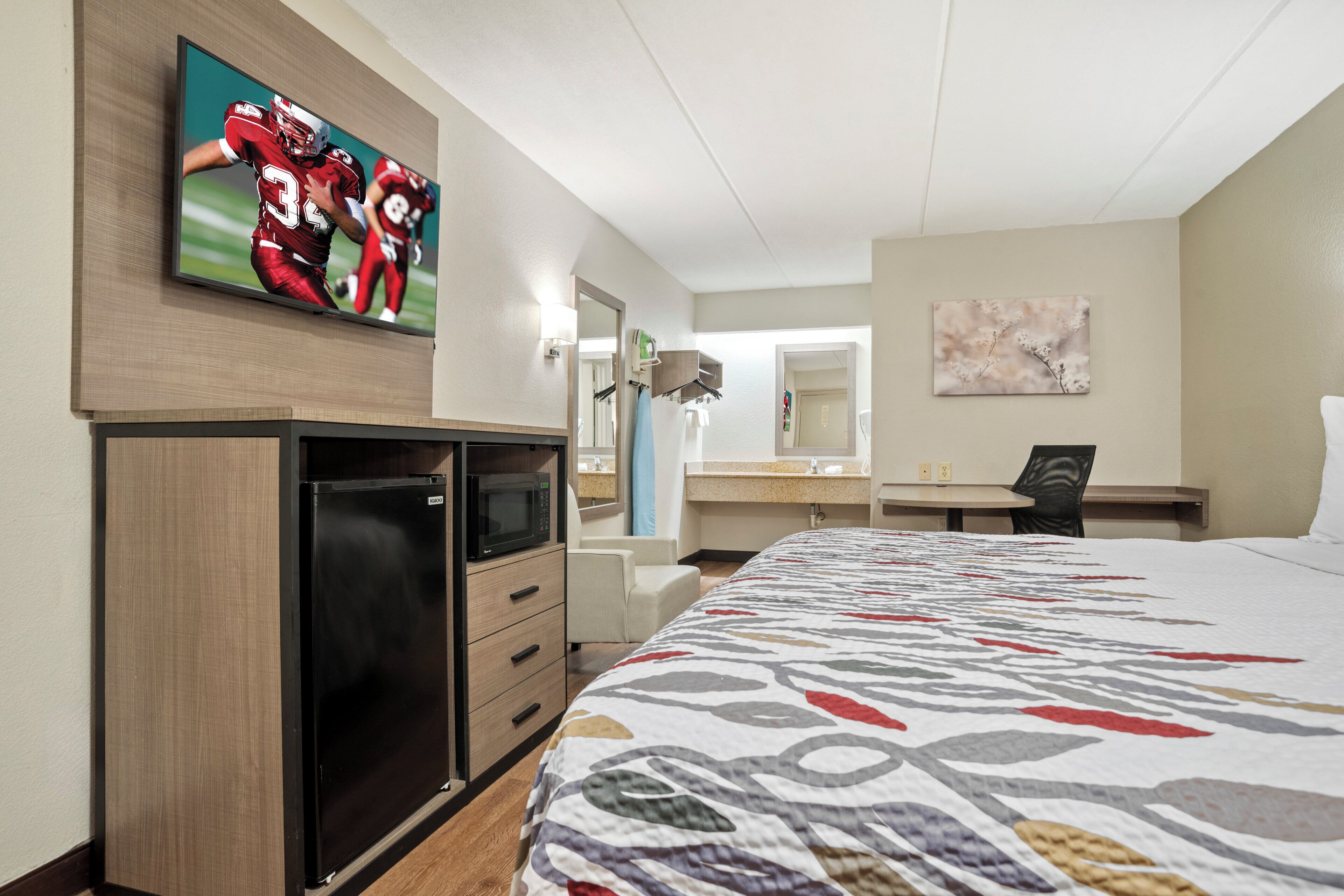 Habitación de hotel con cama, escritorio, televisor montado en la pared, microondas y refrigerador pequeño.