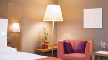 Habitación de hotel con cama, escritorio con lámpara, silla, una pequeña mesa con dos copas de vino y una planta en maceta.