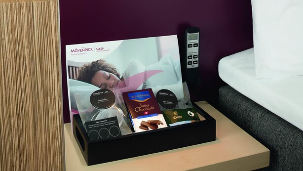 Habitación de hotel con una mesita de noche mostrando una promoción de descanso, un control remoto y una bandeja con una barra de chocolate y antifaz.