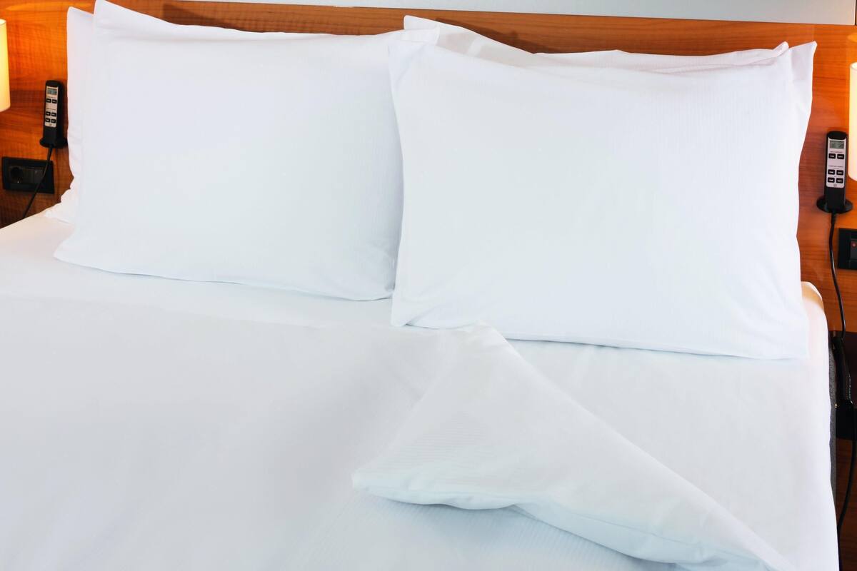 A neatly made bed with white linens and two pillows, a bedside table with a phone and a lamp, and a wooden headboard.