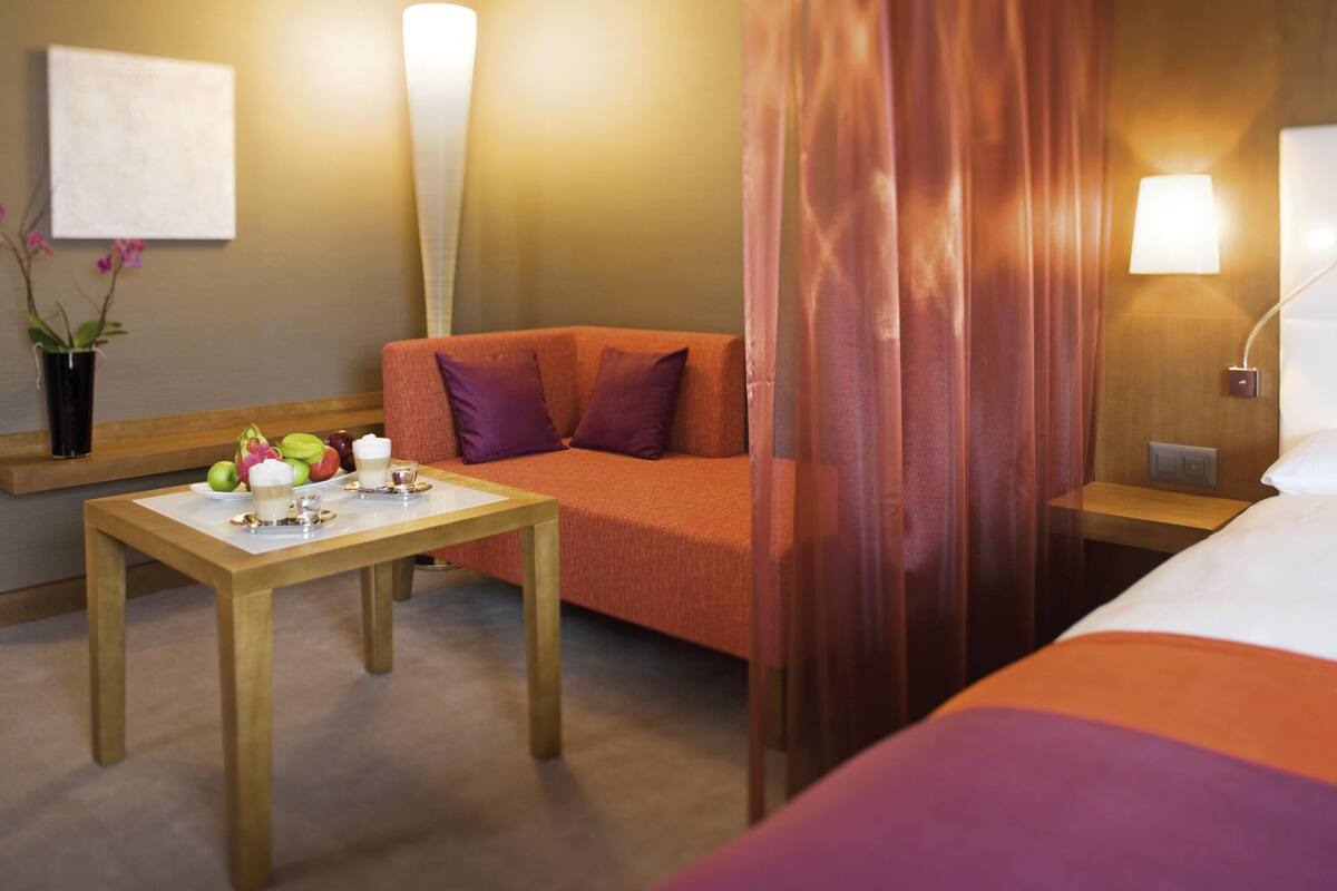 A hotel room with a bed, a sofa, a table with fruits and a drink, and a nightstand with a lamp.