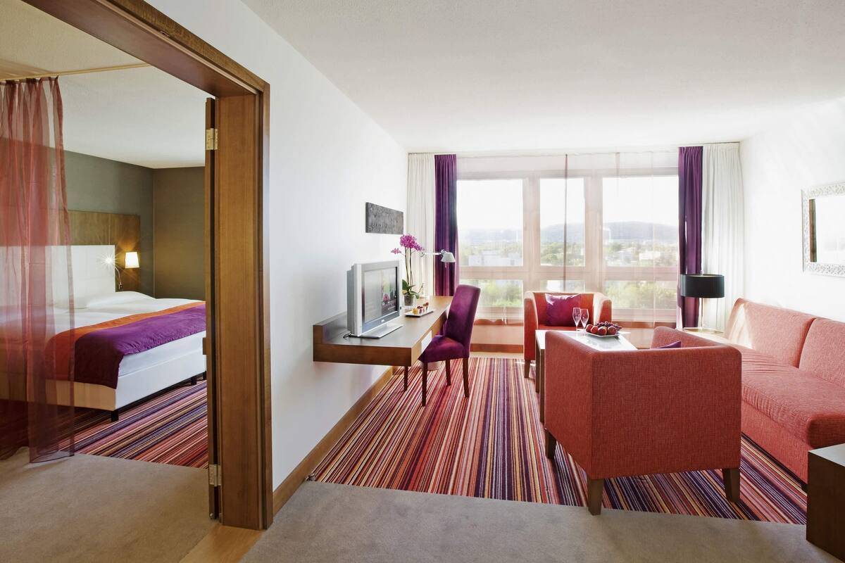 A hotel room with a bed, a desk with a TV, a chair, a sofa, and a view of the city.