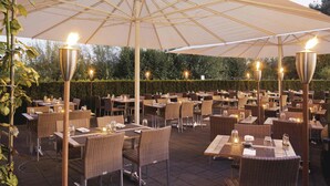 An outdoor dining area with wicker tables and chairs, covered by a large umbrella, and lit by tall, slender lamps. - Mövenpick Hotel Zuerich-Airport (Opfikon)