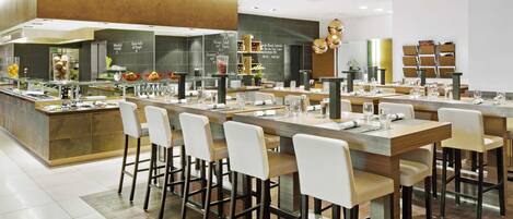 A modern restaurant with a central bar area, multiple seating options, and a visible kitchen in the background.
