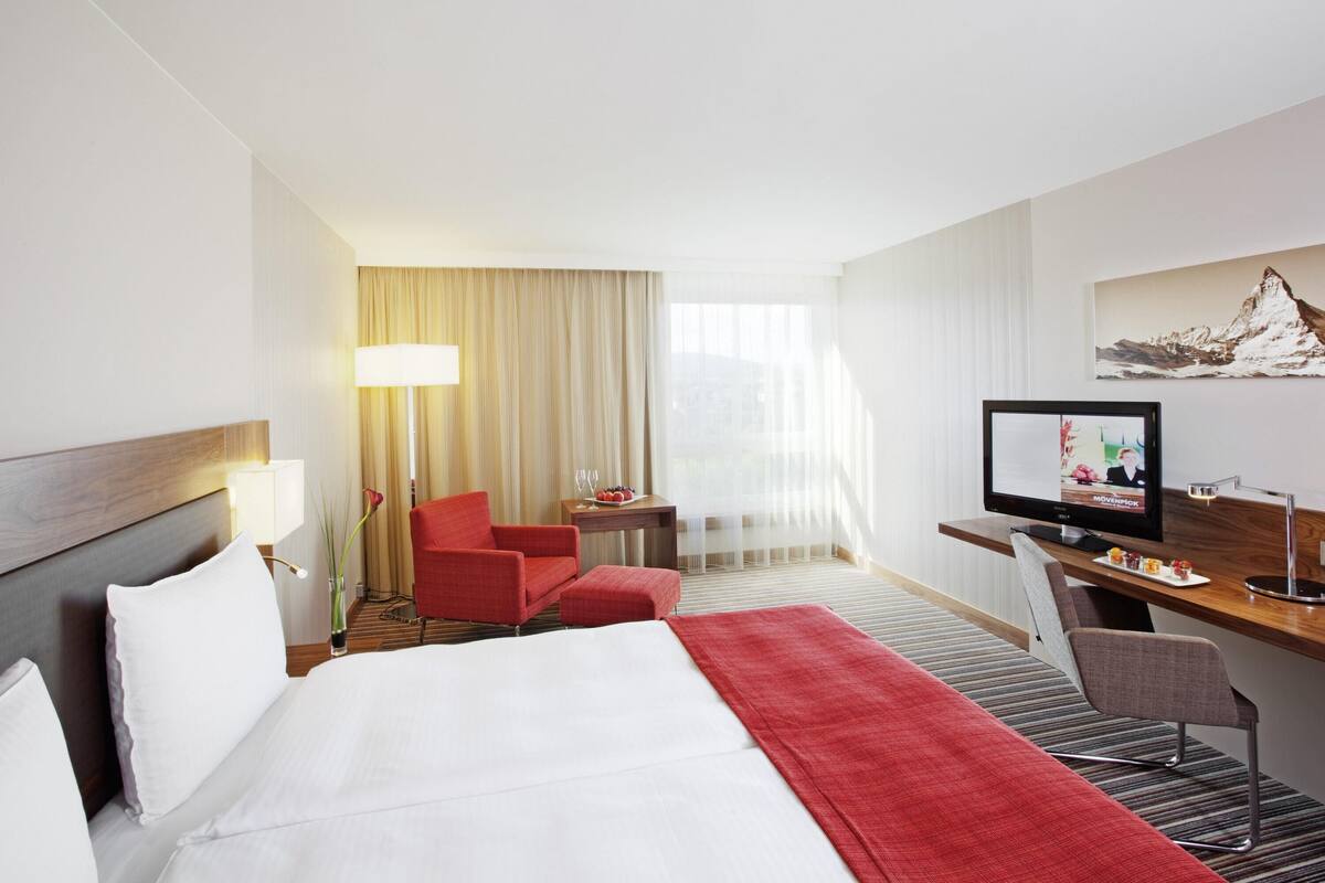 A hotel room with a bed, a desk with a computer, a chair, a lamp, and a window with curtains.