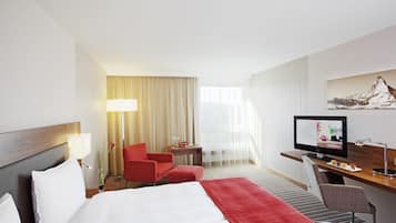 A hotel room with a bed, a desk with a computer, a chair, a lamp, and a window with curtains.
