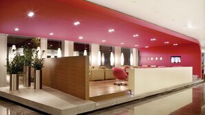 A modern reception area with a red accent wall, a reception desk, seating area, and potted plants. - Mövenpick Hotel Zuerich-Airport (Opfikon)