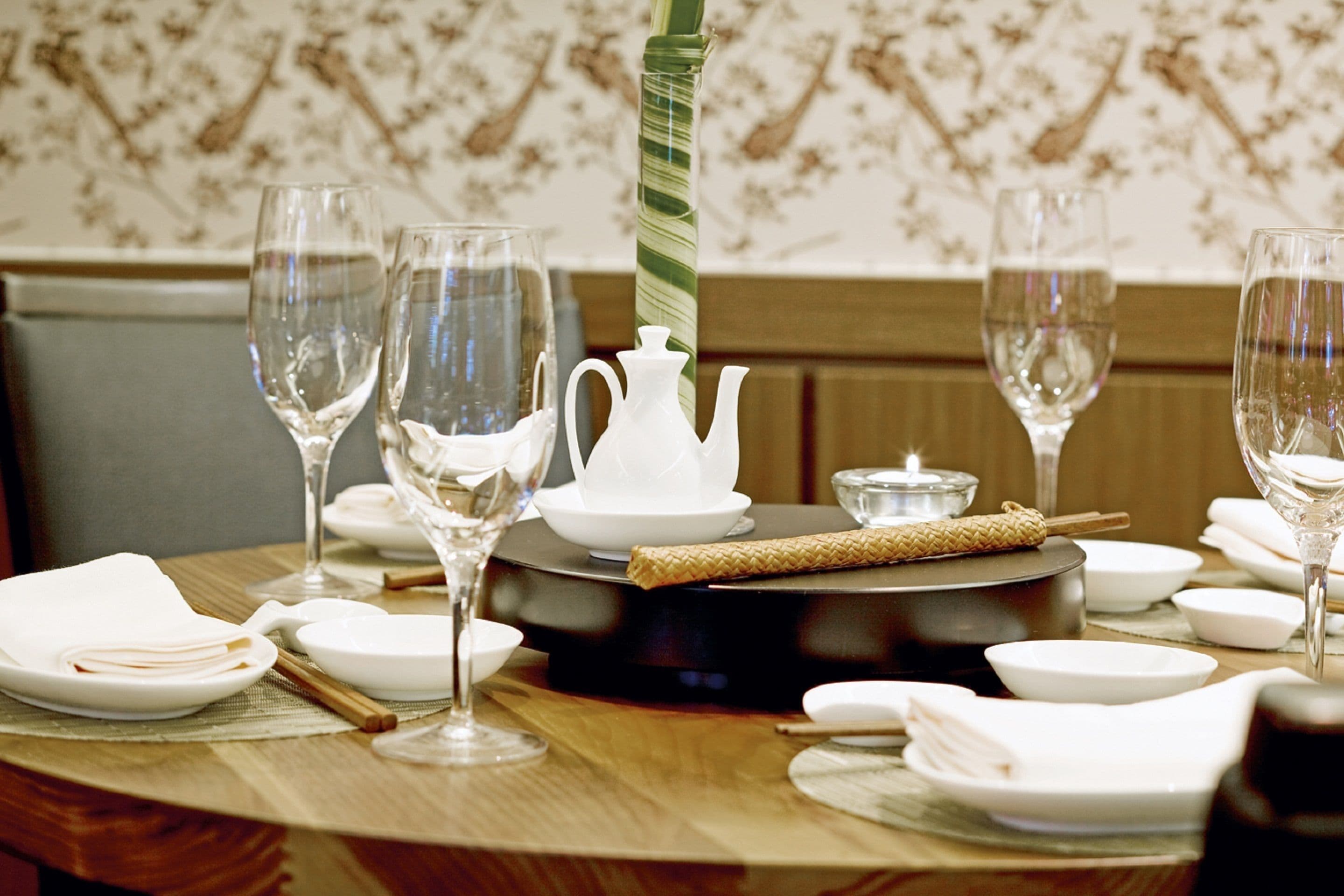 A dining table set with white plates, glasses, and a teapot, with a patterned wallpaper background.