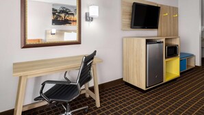 A hotel room with a desk, chair, mirror, television, and built-in storage unit.