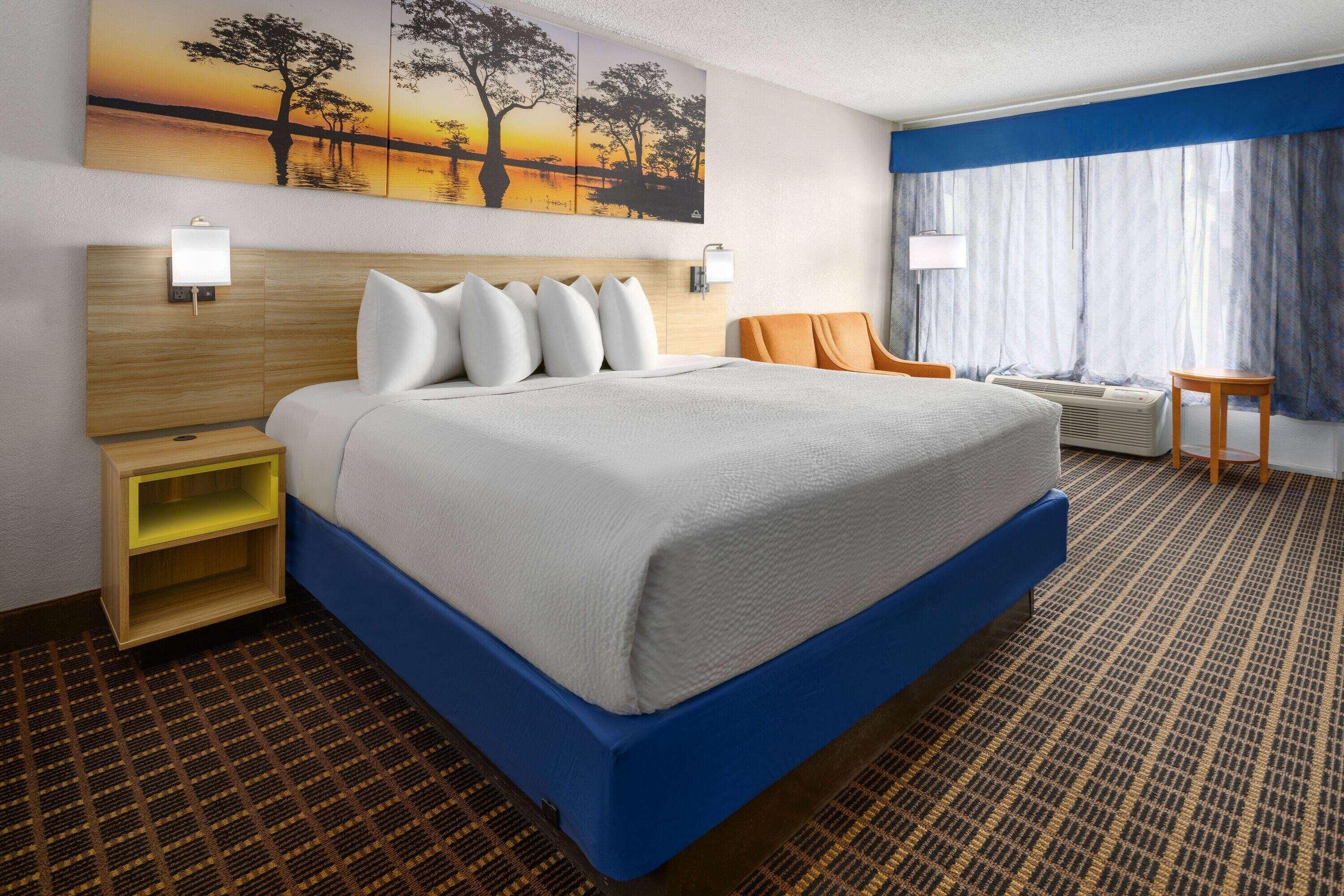 A hotel room with a large bed, a nightstand, a desk, a chair, a window with curtains, and a wall painting.