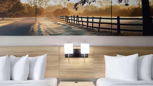 A hotel room with a large wall mural, a wooden floor, a bed with white bedding, two bedside tables with lamps, and a small yellow nightstand.