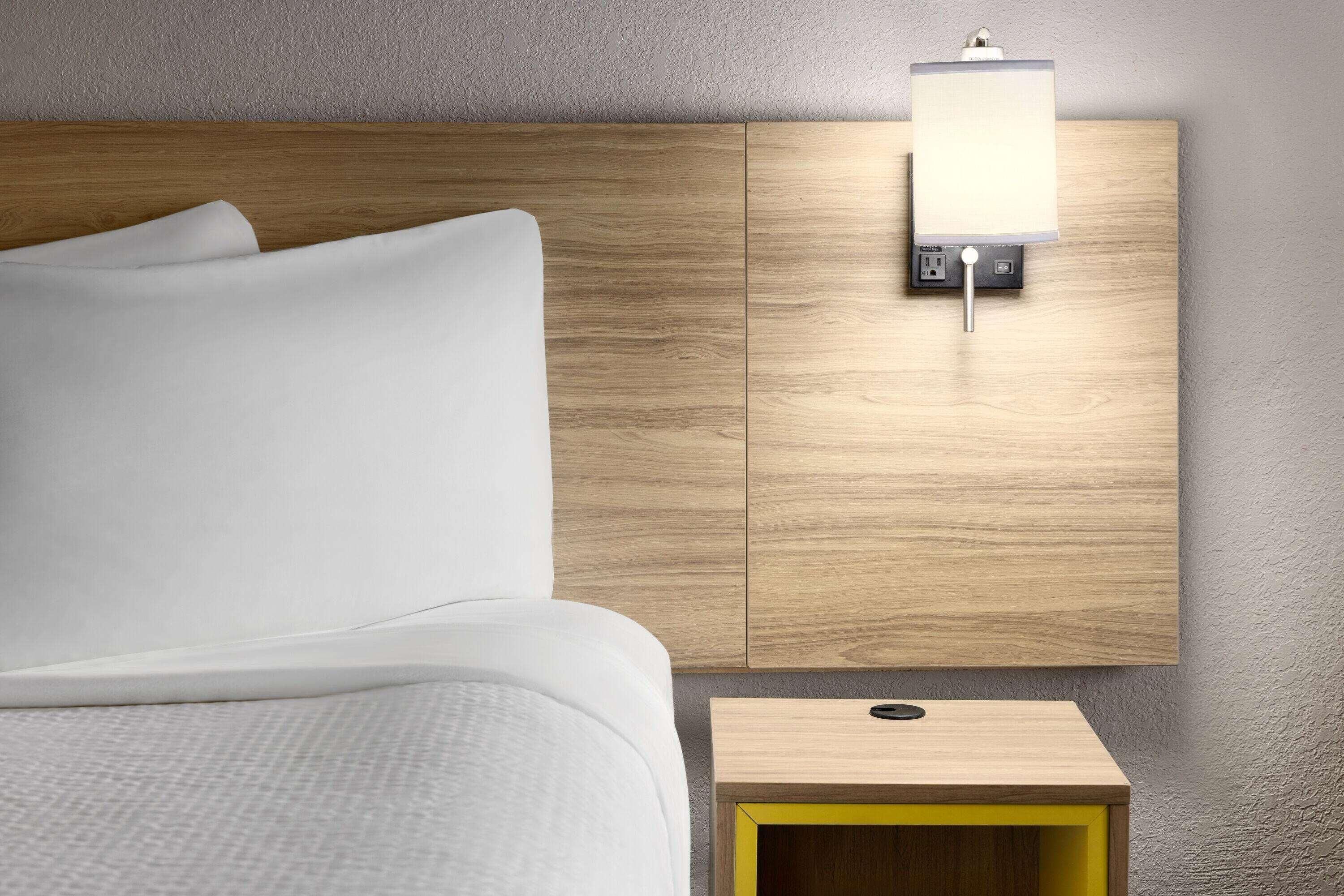 A bed with white linens, a wooden headboard, a bedside table with a yellow accent, and a wall-mounted light fixture.