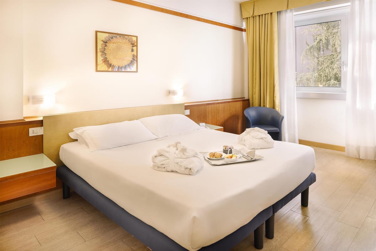 Une chambre d’hôtel avec un grand lit, une tête de lit, une table de chevet, une chaise, une fenêtre avec des rideaux et une œuvre d’art accrochée au mur.