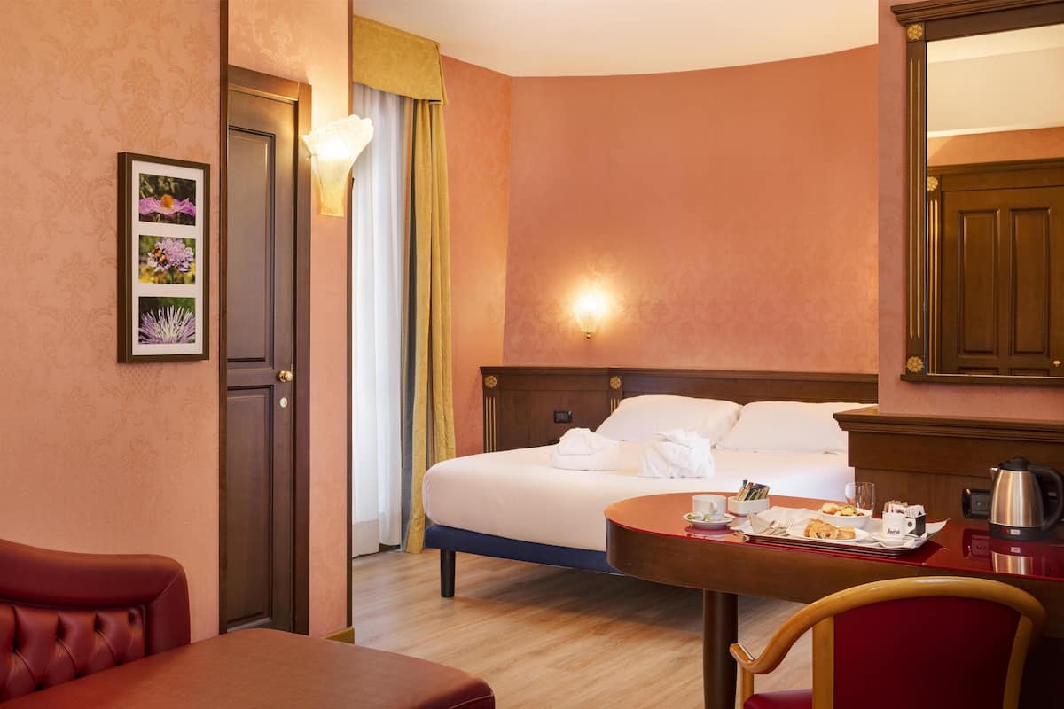 Une chambre d’hôtel avec un lit, une table à manger avec des aliments pour le petit-déjeuner, une chaise, un canapé rouge, un tableau encadré au mur et une porte.