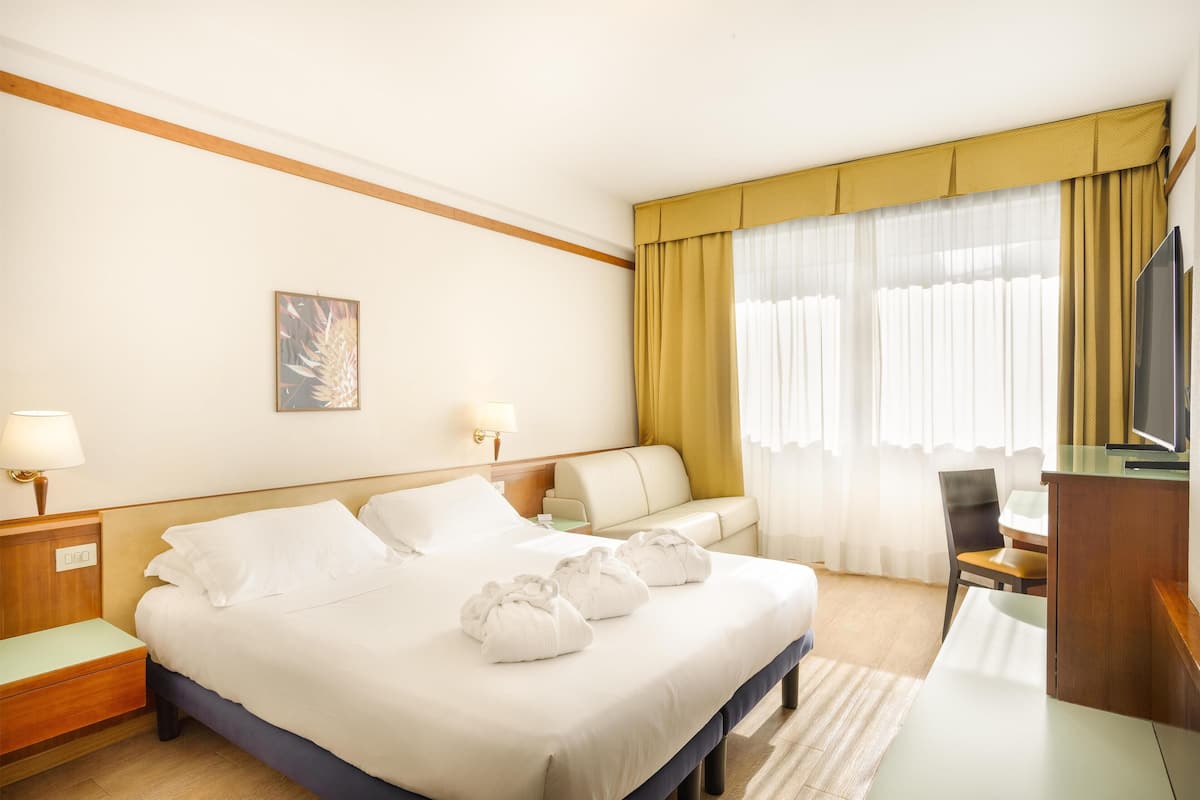 Une chambre d’hôtel avec un lit, un bureau, une chaise, une télévision et une fenêtre avec des rideaux.