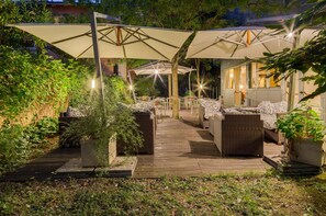 An outdoor seating area with wicker furniture and umbrellas, surrounded by greenery and a wooden deck.