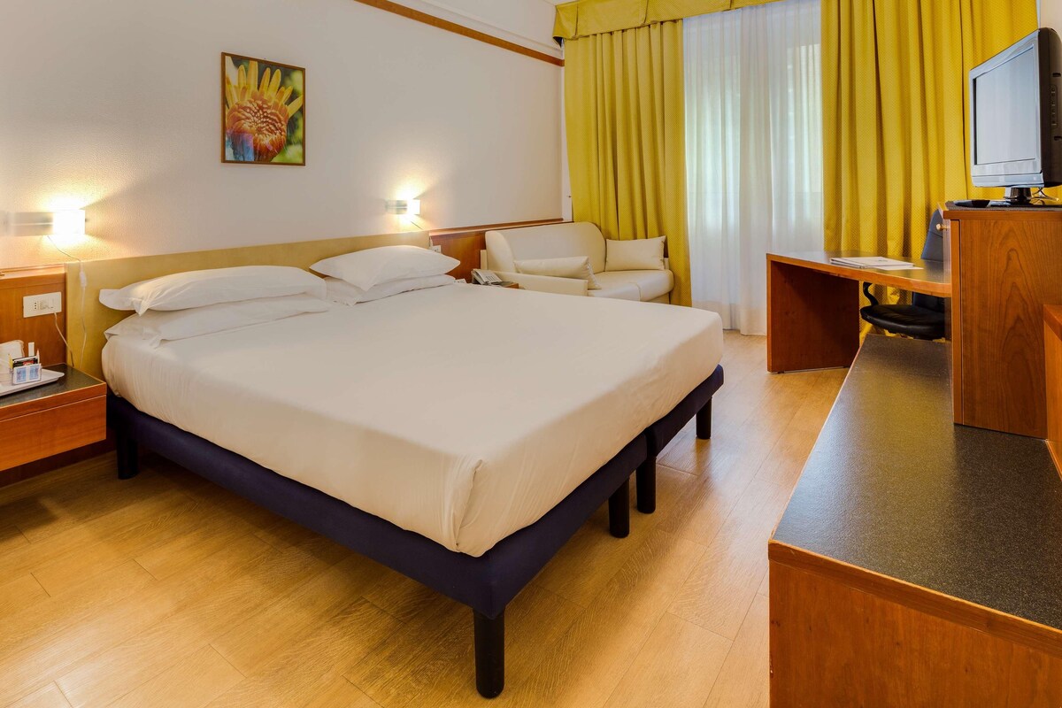 Ein Hotelzimmer mit einem großen Bett, Nachttischen, einem Schreibtisch, einem Sessel, einem Fernseher und einem Bild an der Wand.