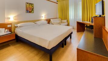 Ein Hotelzimmer mit einem großen Bett, einem Schreibtisch mit Computer, einem Sessel und einem kleinen Tisch mit Telefon.